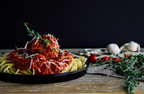 A plate of spaghetti topped with marinara sauce, herbs, and grated cheese, with garlic and tomatoes in the background.