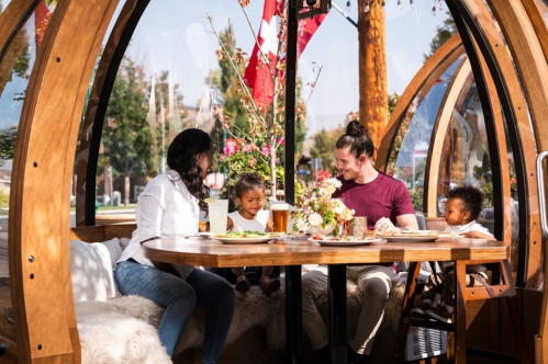 A family enjoys a meal in a cozy, modern dining pod, surrounded by greenery and flags. Two children are present.