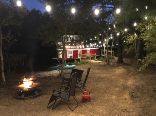 A cozy outdoor scene with a fire pit, chairs, and string lights illuminating a red building in the background.