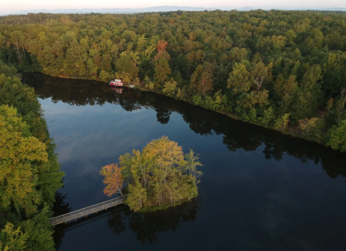 Aerial view of a serene lake surrounded by lush trees, featuring a small island and a boat in the distance.