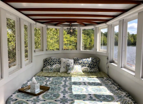 Cozy interior of a boat with a bed, decorative pillows, and large windows overlooking a serene lake.