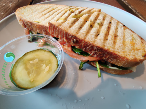 A grilled sandwich with tomatoes and greens, served with a side of pickles and a small container of sauce.