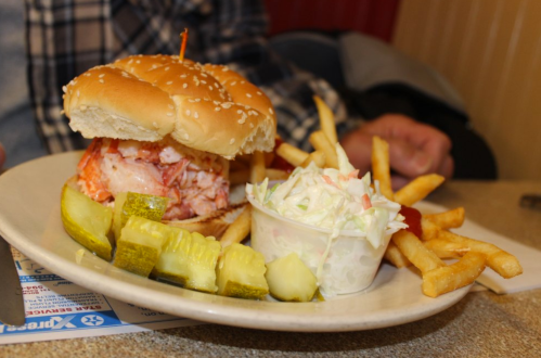 A plate with a lobster sandwich, fries, coleslaw, pickles, and ketchup on a table.
