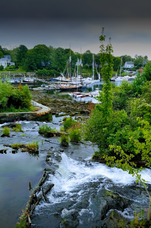 A serene harbor scene with boats docked, surrounded by lush greenery and a flowing stream in the foreground.