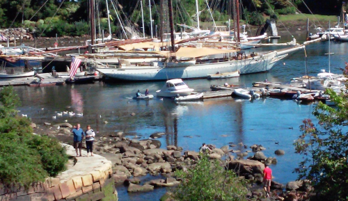 A scenic harbor with boats docked, people walking on rocks, and lush greenery in the background.