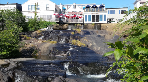 A restaurant overlooks a rocky waterfall, surrounded by greenery and blue skies. Red umbrellas are visible on the patio.