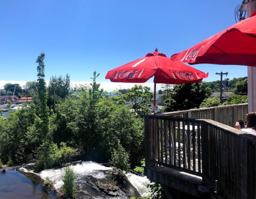 A sunny outdoor patio with red umbrellas overlooking a river and boats in the distance, surrounded by greenery.