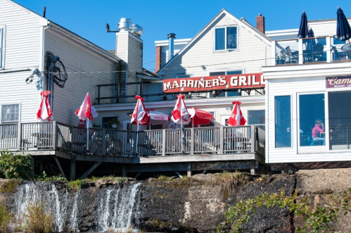 A waterfront restaurant with outdoor seating and red umbrellas, featuring a sign that reads "Marriner's Grill."