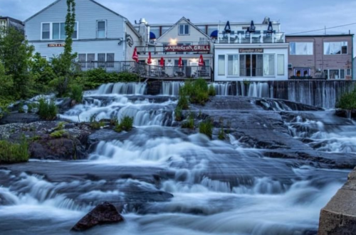 A serene waterfall flows over rocks, with a restaurant in the background under a cloudy sky.