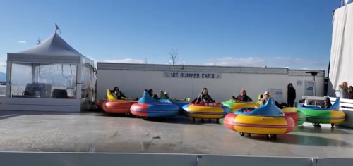 A row of colorful bumper cars on an ice rink, with people seated inside, under a clear blue sky.