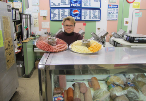 A woman stands behind a deli counter, holding two platters of sliced meats and cheeses, with various products displayed below.