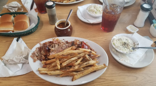 A plate of pulled pork with barbecue sauce, seasoned fries, and sides of coleslaw and chili on a wooden table.