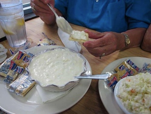 A person holds a cracker topped with creamy dip, with a bowl of dip and a side of coleslaw on the table.