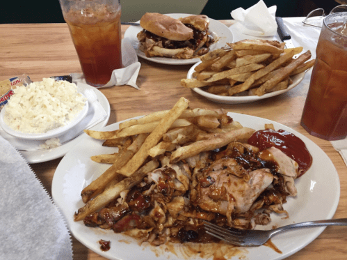 A plate of BBQ chicken with fries and coleslaw, alongside a sandwich and iced tea on a wooden table.