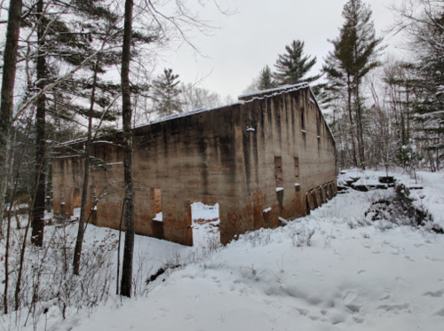 Abandoned building partially covered in snow, surrounded by trees in a winter landscape.