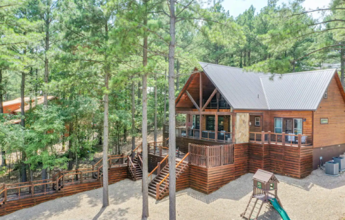 A wooden cabin surrounded by tall trees, featuring a deck and playground in a serene outdoor setting.