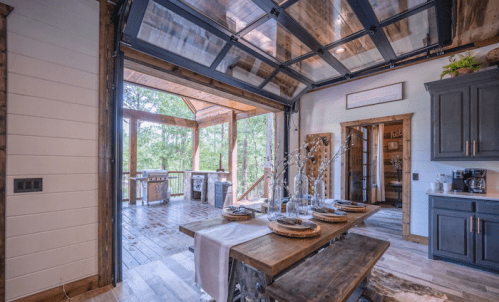 A rustic kitchen with a wooden dining table, open to a deck through a large glass garage door, surrounded by trees.