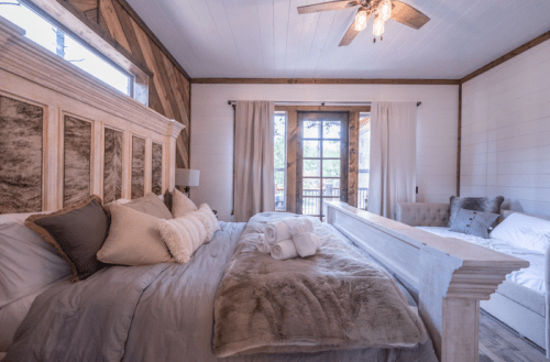 Cozy bedroom with a large bed, soft linens, and natural light from windows, featuring rustic decor and a plush armchair.