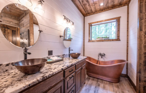 A rustic bathroom featuring dual round mirrors, a wooden vanity with copper sinks, and a freestanding copper bathtub.