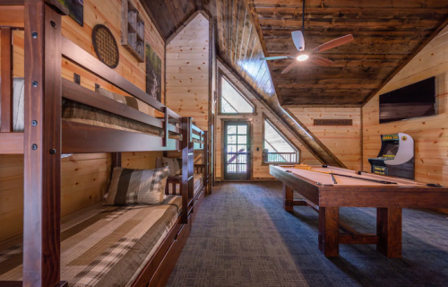 Cozy cabin interior with bunk beds, a pool table, and large windows overlooking a wooded area.