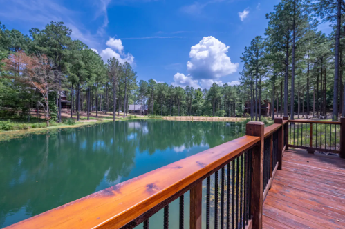 A serene pond surrounded by tall pine trees under a blue sky with fluffy clouds, viewed from a wooden deck.