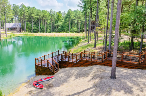 A serene pond surrounded by trees, featuring a wooden deck and two kayaks on the shore.