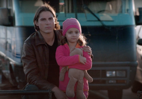 A man and a young girl sit together, the girl holding a stuffed toy, with a truck in the background.