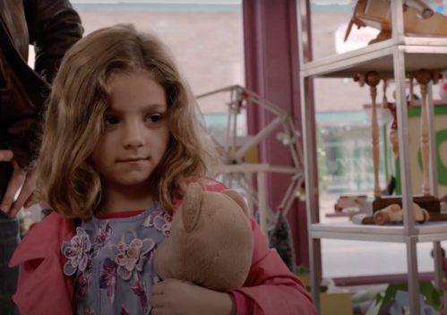 A young girl with wavy hair holds a stuffed animal, looking thoughtfully in a colorful toy store.