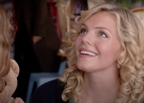 A smiling young woman with curly blonde hair looks up, engaged in a conversation, in a lively setting.