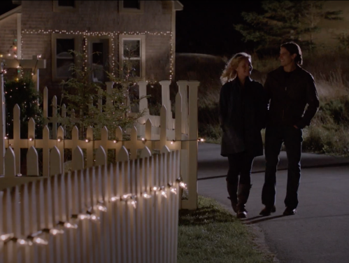 A couple walks together along a lit path, surrounded by festive decorations and a cozy house at night.