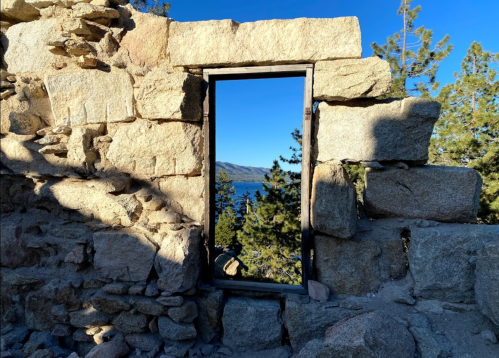 A stone wall with a window frame, overlooking a lake and trees in the background under a clear blue sky.