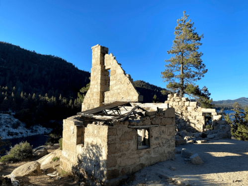 Ruins of a stone building with a collapsed roof, surrounded by trees and mountains under a clear blue sky.