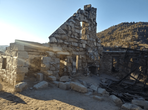 Ruins of a stone building with a partially collapsed wall, surrounded by trees and mountains under a clear blue sky.