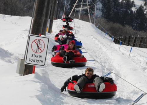 A line of people on snow tubes being pulled up a snowy hill, with a "Stay in Tow Path" sign nearby.