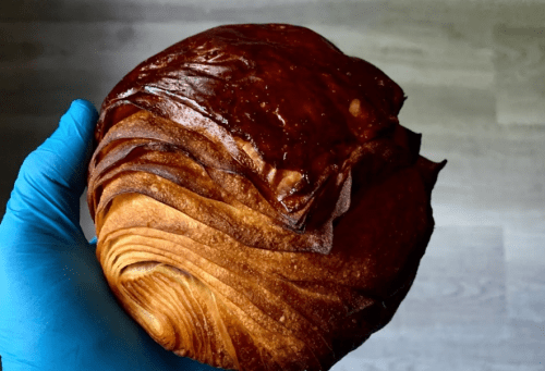 A hand in a blue glove holds a glossy, layered pastry with a golden-brown crust.