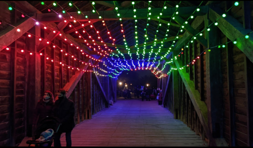 A wooden bridge adorned with colorful string lights in red, green, and blue, with people walking underneath.