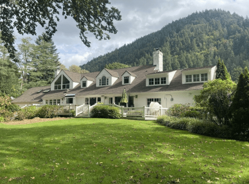 A large white house with a sloped roof, surrounded by greenery and mountains in the background under a cloudy sky.