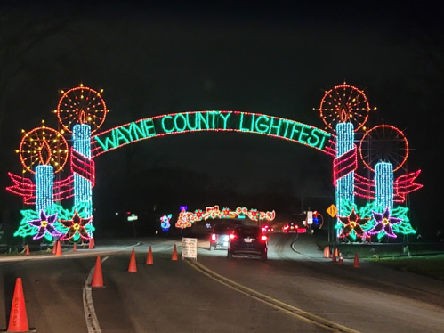 Colorful holiday lights arch over a road, reading "Wayne County Lightfest," with festive decorations and traffic cones.