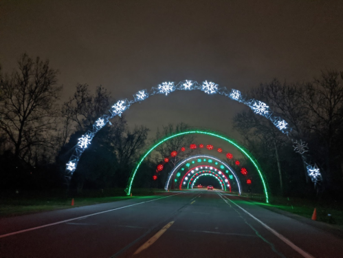 Colorful holiday light arches with snowflakes and red accents illuminate a dark road, creating a festive atmosphere.
