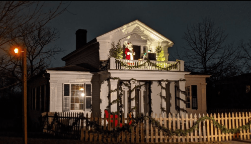 A decorated house at night with Santa on the balcony, surrounded by lights and greenery, and a picket fence.