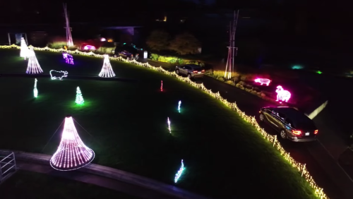 Aerial view of a festive display with colorful lights, including trees and animals, alongside a winding driveway with cars.