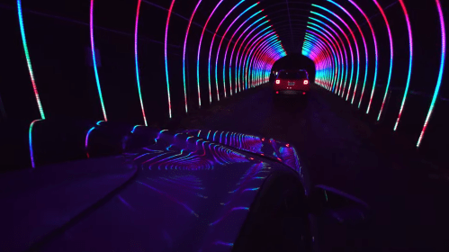 A car drives through a colorful, illuminated tunnel with vibrant neon lights creating a dynamic pattern.