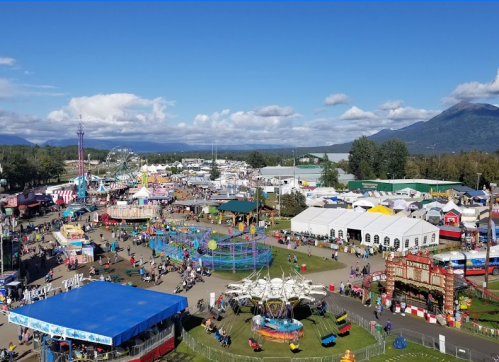 Aerial view of a vibrant fairground with rides, games, and crowds, set against a backdrop of mountains and blue skies.