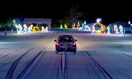 A car drives down a snowy road, passing colorful holiday lights and decorations at night.