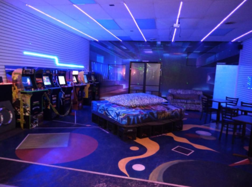A retro arcade room with neon lights, arcade machines, a cozy seating area, and colorful floor designs.