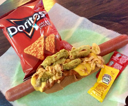 A hot dog topped with cheese, fried onions, and jalapeños, served with a side of Doritos and ketchup packets.