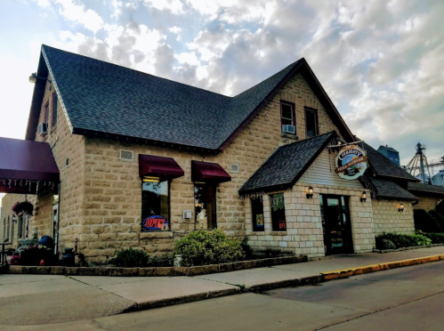 A stone building with a sloped roof, featuring red awnings and an "Open" sign, set against a cloudy sky.