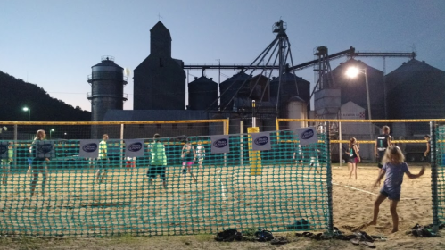 A beach volleyball game at dusk near industrial silos, with players and spectators enjoying the evening.