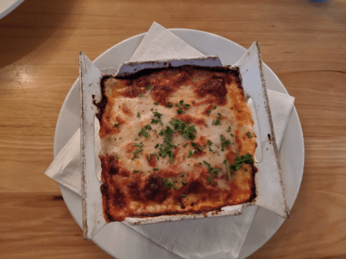 A baked lasagna in a white dish, topped with melted cheese and garnished with parsley, served on a wooden table.