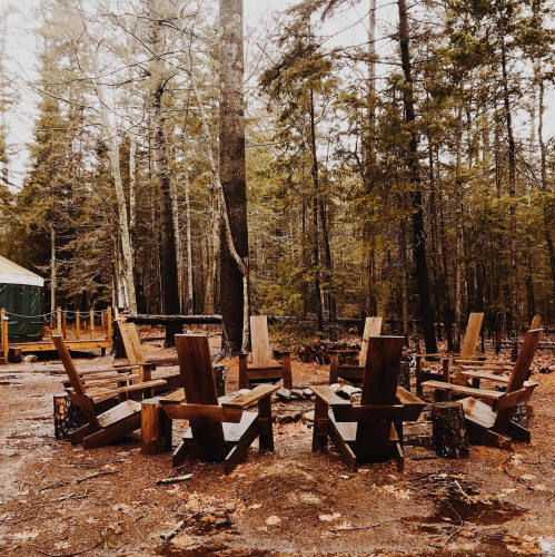 A circle of wooden chairs around a fire pit in a forested area, surrounded by tall trees and a misty atmosphere.